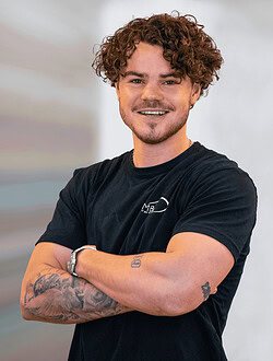 Mand med krøllet hår og tatoveringer står med armene over kors